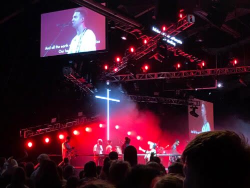 Live Christian worship band performing on stage with a large illuminated cross and red lighting.